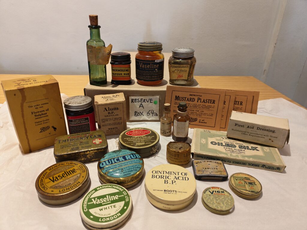 Multiple old fashioned medicine tins, bottles and packets arranged around a brown box labelled 'Reserve A, Box 64a' The collection includes 7 jars and bottles, 9 tins, 4 card boxes and 4 flat packets ladled Mustard Plaster. Visible labels show products such as 'vaseline, ointment of Boric acid BP, first aid dressing, quick rub, emergency case, oiled silk.