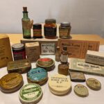 Multiple old fashioned medicine tins, bottles and packets arranged around a brown box labelled 'Reserve A, Box 64a' The collection includes 7 jars and bottles, 9 tins, 4 card boxes and 4 flat packets ladled Mustard Plaster. Visible labels show products such as 'vaseline, ointment of Boric acid BP, first aid dressing, quick rub, emergency case, oiled silk.