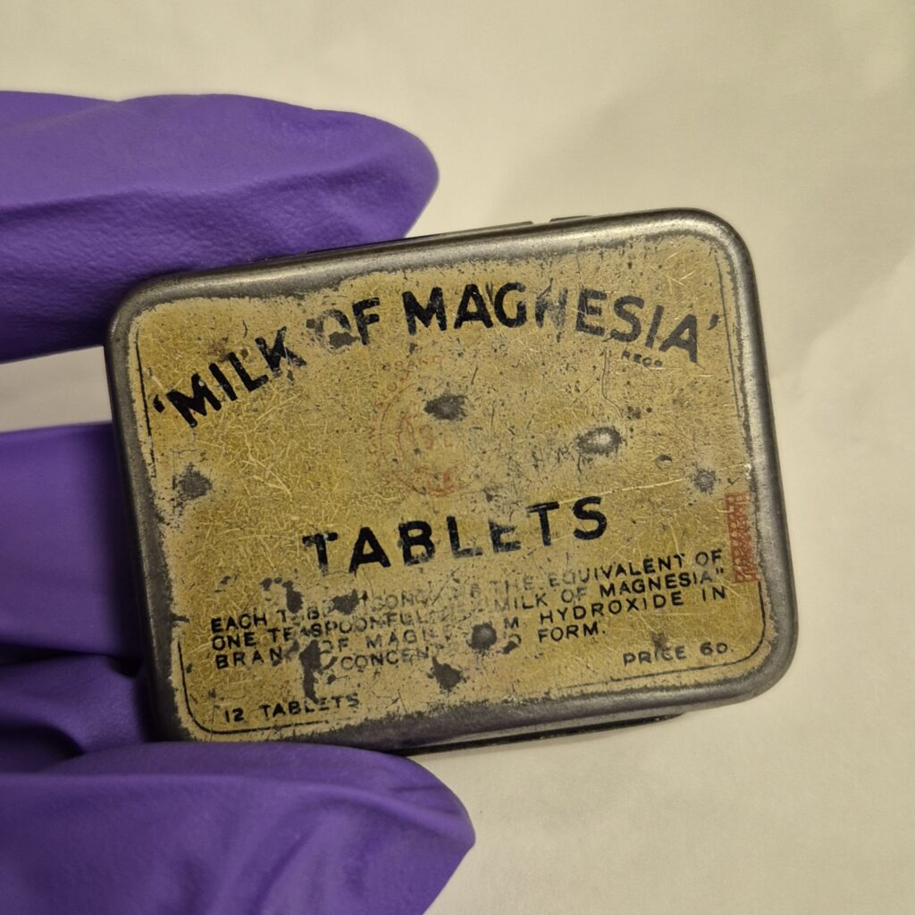 Small rectangular tin made of metal. A cream label reads 'Milk of Magnesia tablets' in Black writing.