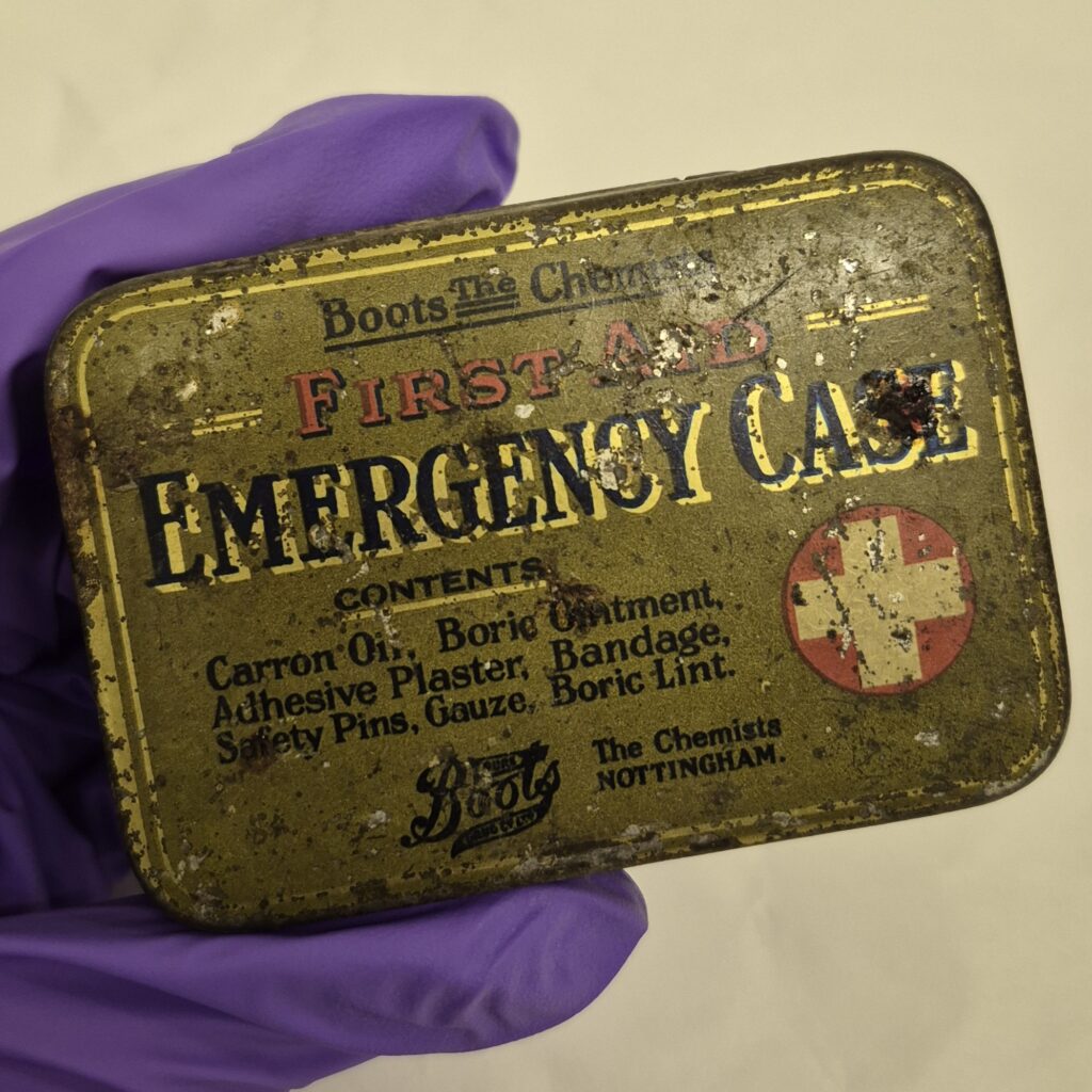 Metal rectangular green tin with worn and rusted lid. Text in black and red reads 'Boots the Chemists, First Aid, Emergency Case'. Also features the boots logo and first aid symbol of a white cross in a red circle.