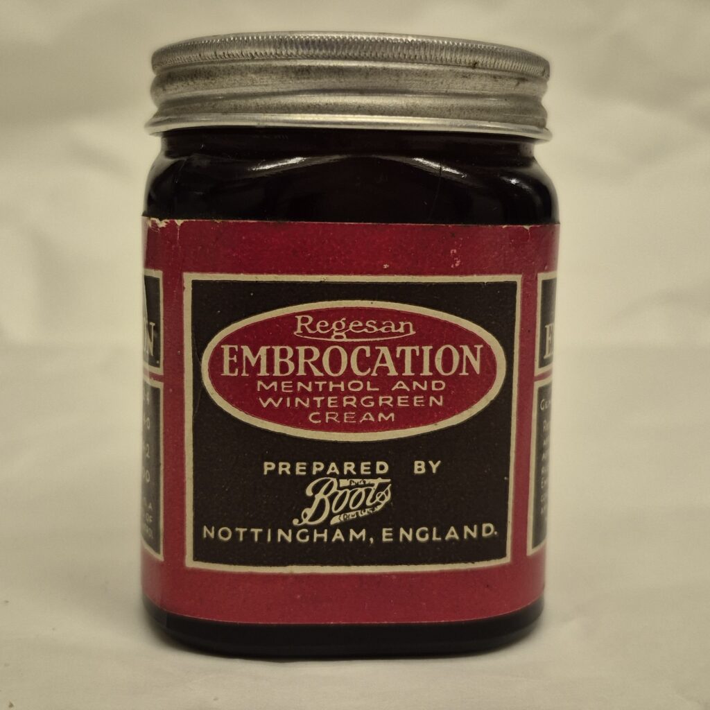 Square jar of black glass with metallic lid. Red label features black box of text. The box includes a red oval that in cream text reads; 'Regesan, Embrocation Methonal and Wintergreen Cream' Underneath the text reads 'Boots, Nottingham.'