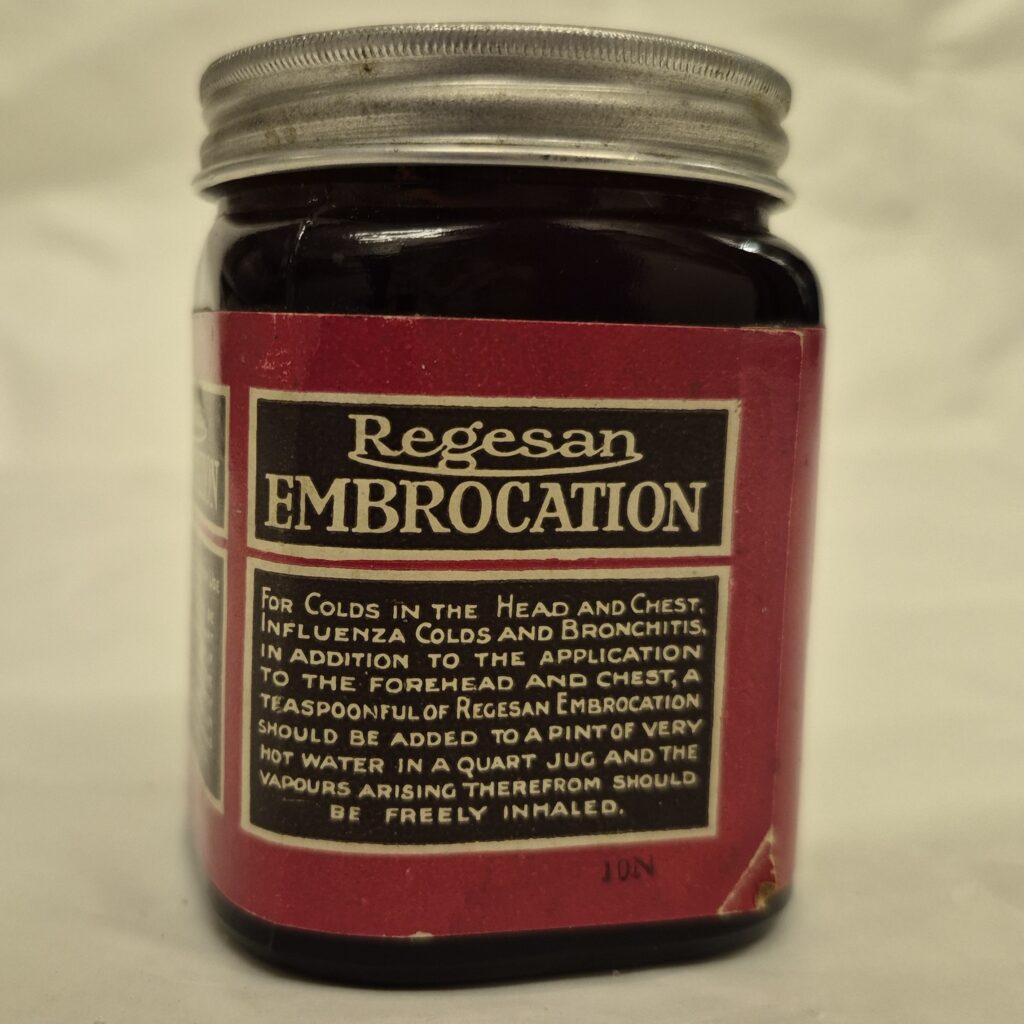 Square jar of black glass with metallic lid. Red label features two black boxes of cream text. The first box reads, Regesan Embrocation and the bottom reads with directions of use.