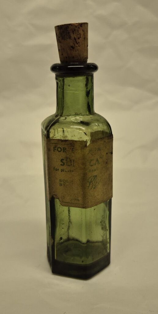 Green hexagonal bottle with cork stopper. A faded and worn label features mostly illegible text.