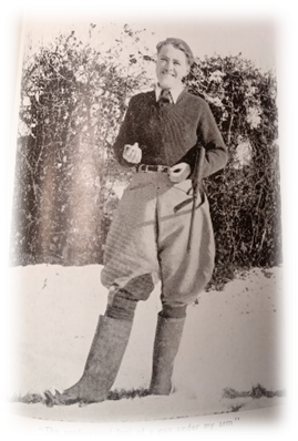 Black and White image of WW2 land-girl wearing a shirt, tie, heavy woollen jumper, plus fours and tall boots. A shot gun hangs under her arm.