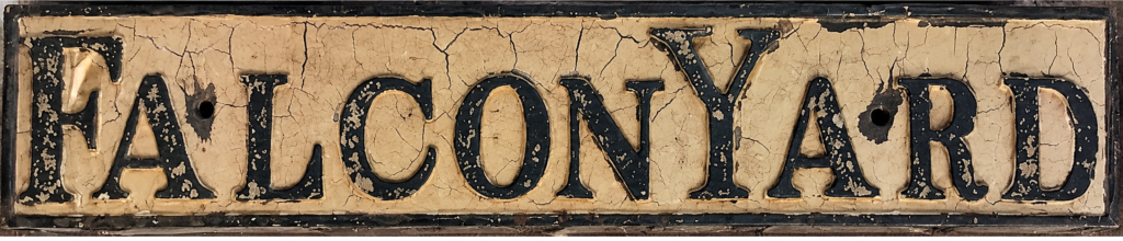 Cream street sign with black capitalised text which reads Falcon Yard. paint is cracked and worn.
