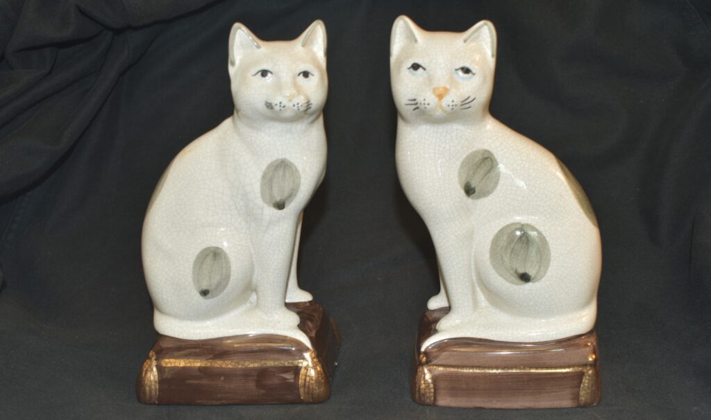 Two ceramic, white cat statuettes with painted features including spots on bodies. Each cat sits on a brown cushion; bodies facing each other with their face looking toward the viewer.