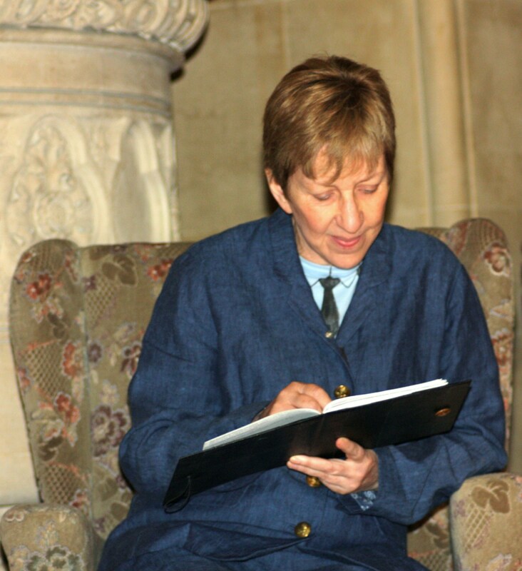 Colour photograph of white woman with short light brown hair sitting in an armchair reading a book. She is wearing a blue suit with a shirt and tie.
