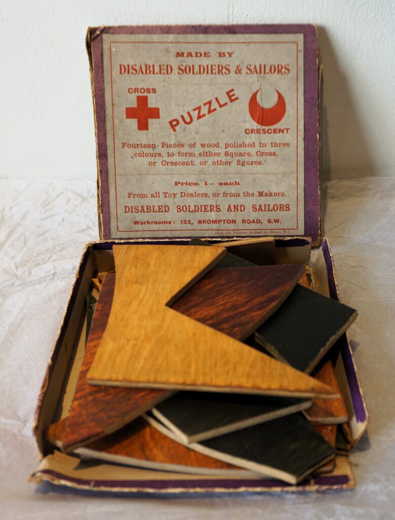 Square box of geometric puzzle pieces with the lid of the box propped up behind it. The box lid is mainly cream with a purple border, red text reads 'Made by Disabled Soldiers & Sailers' under this there is a symbol of a cross on the right, and a crescent shape on the left, in the middle the word 'PUZZLE' is written in diagonal text. Below this as smaller print text of instructions and details of the puzzles manufacture.
In the box wooden pieces are stacked, the top piece is yellow and cut into a chevron shape. The pieces below are of yellow, red and green.