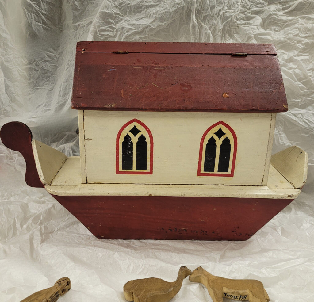 Wooden toy in the shape of a boat (Noah's Ark). The base and roof of the ship is a burgundy red and there is a glimpse of hinges near the top of the roof that suggests that the roof opens up. The rest of the Ark is cream, and the main part has two painted arched windows with black windowpanes and outlined in red.