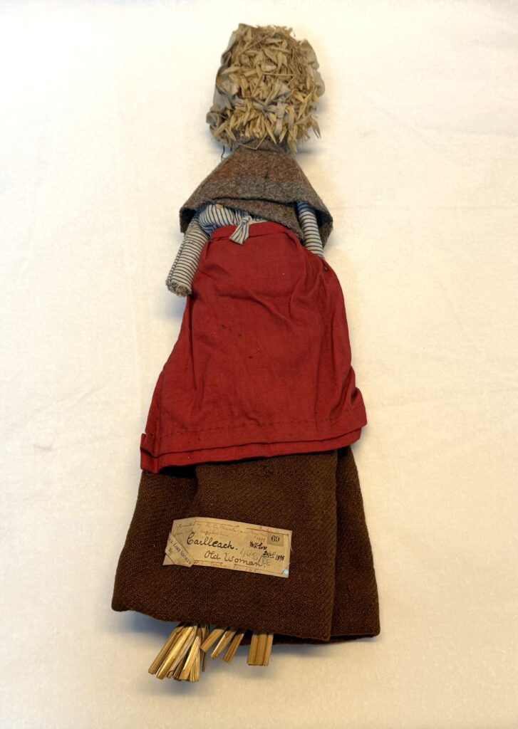 Soft doll with a corn tassel face and corn stalk feet. It is clothed in a white bonnet, brown woollen shawl, blue and white striped shirt, red apron and brown skirt.