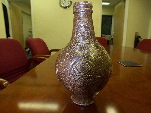Brown bottle sitting on a corporate meeting table. The bottle is terracotta coloured with a white shine which highlights a abstract face on the neck and a daisy wheel on the round body.