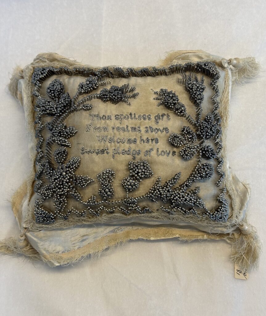 Rectangular, cream, silk cushion with cream tassels and fringe. Hundreds of steel pins are placed in a three-dimensional floral pattern and boarder. In the centre a message is speeled out using smaller pins. The message reads ‘Thou spotless gift, from reeling above, Welcome here sweet pledge of love.’