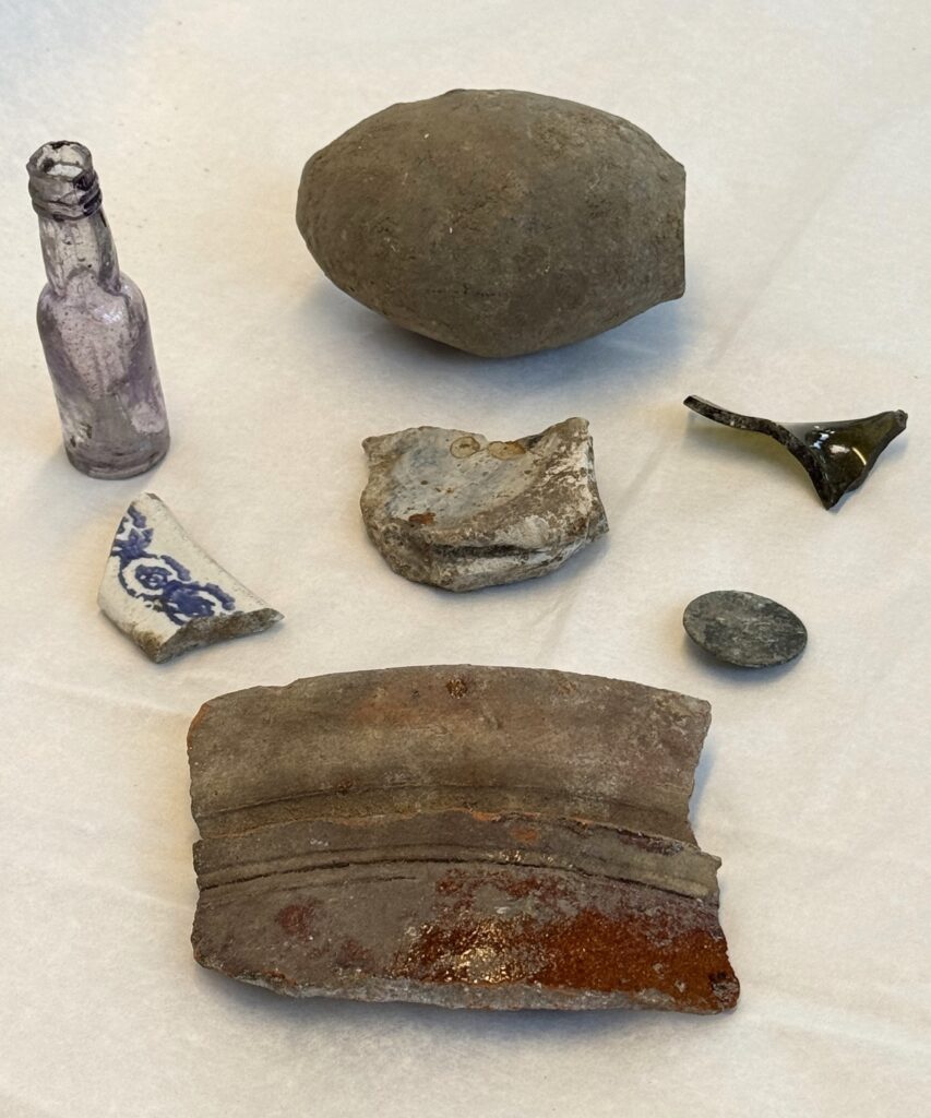 Seven objects displayed in two rows of three and one larger piece at the bottom. Moving lert to right from top to bottom; a small, lilac tinted glass bottle, an egg shaped, smooth grey stone, a dark grey/ green shard of bent glass. Second row; A broken piece of pottery with white glaze painted with a blue circular pattern, a piece of unglazed grey pottery, tarnished metallic button which is very eroded. On the bottom is a larger piece of terracotta pot.