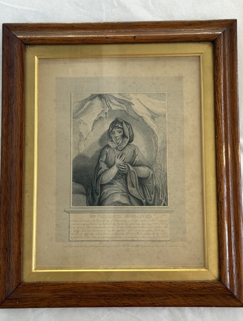 Greyscale etching in a brown frame with a gold inner frame. The etching shows a woman, Elizabeth Woodcock, in a snow cave. She is wearing a flowing, hooded cape. Her hands are held at her chest. Beneath this a square of had written text is headed Mrs. Elizabrth Woodcock with the story of her ordeal in smaller handwriting.