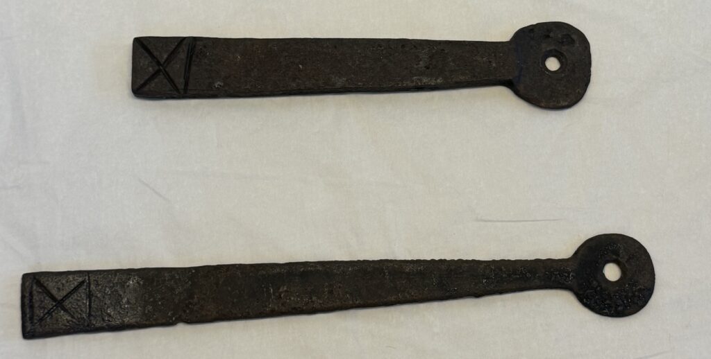 Two black metal door latches both an elongated keyhole shape with a hole in the bulbous end. Both feature a engraved mark at the squared end in the shape of an X with a line above and below. The latch that is displayed above is shorter than the one below.