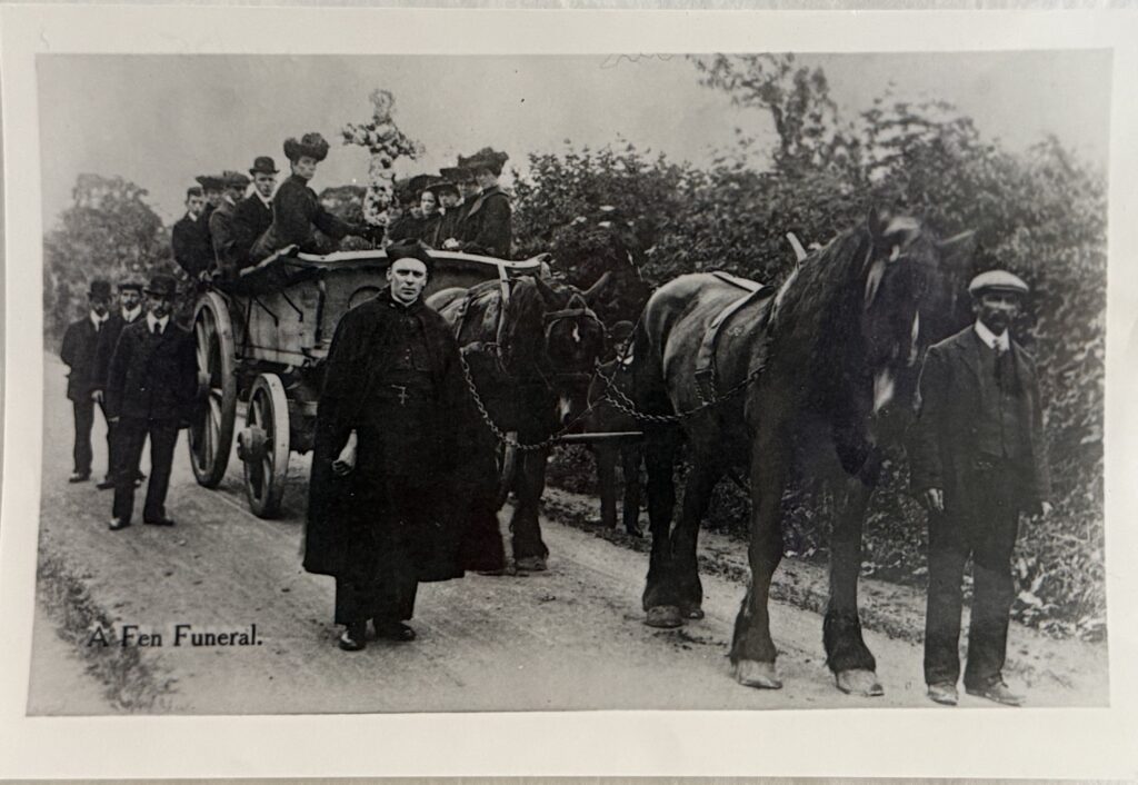 Black and white photograph of six figures standing around the front end of a cart pulled by two horses upon which men and women are seated. The figures are wearing period dress from around the 1890s including bowler hats and flat caps. a priest in a cloak and square hat is in the centre. On the cart is a floral cross.