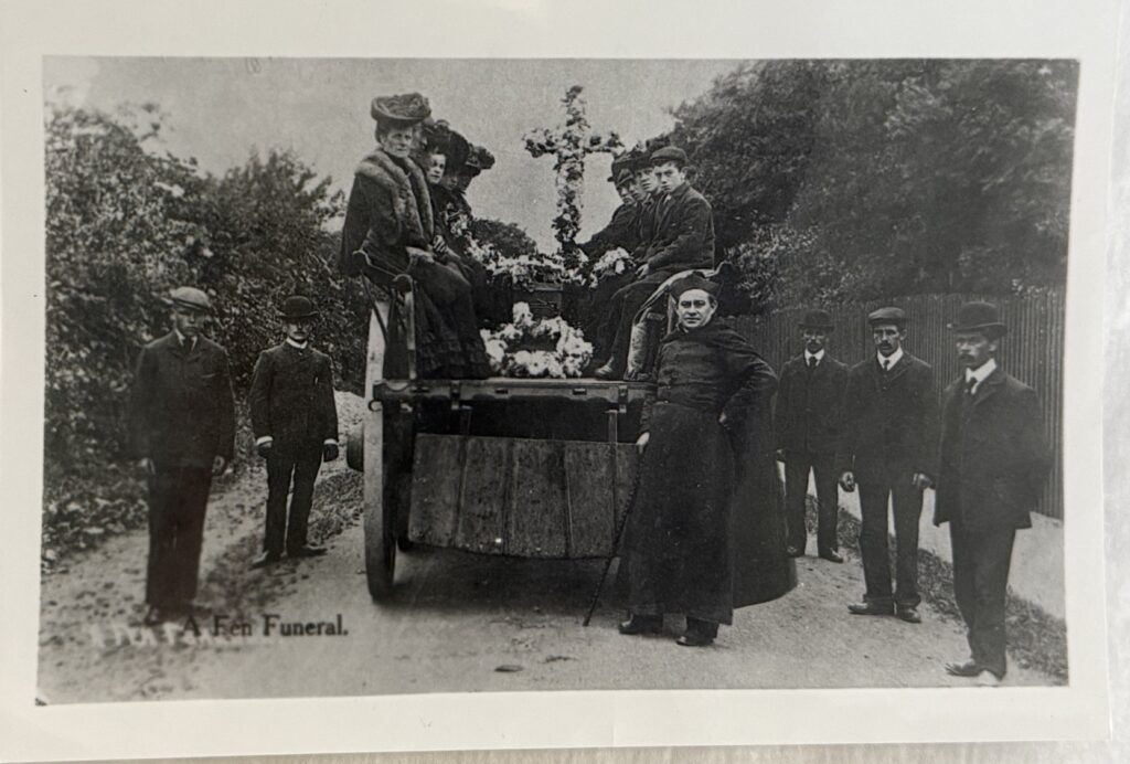 Black and white photograph of six figures standing around the back end of a cart upon which men and women are seated. The figures are wearing period dress from around the 1890s including bowler hats and flat caps. a priest in a cloak and square hat is in the fore. On the cart is a floral wreath and cross.