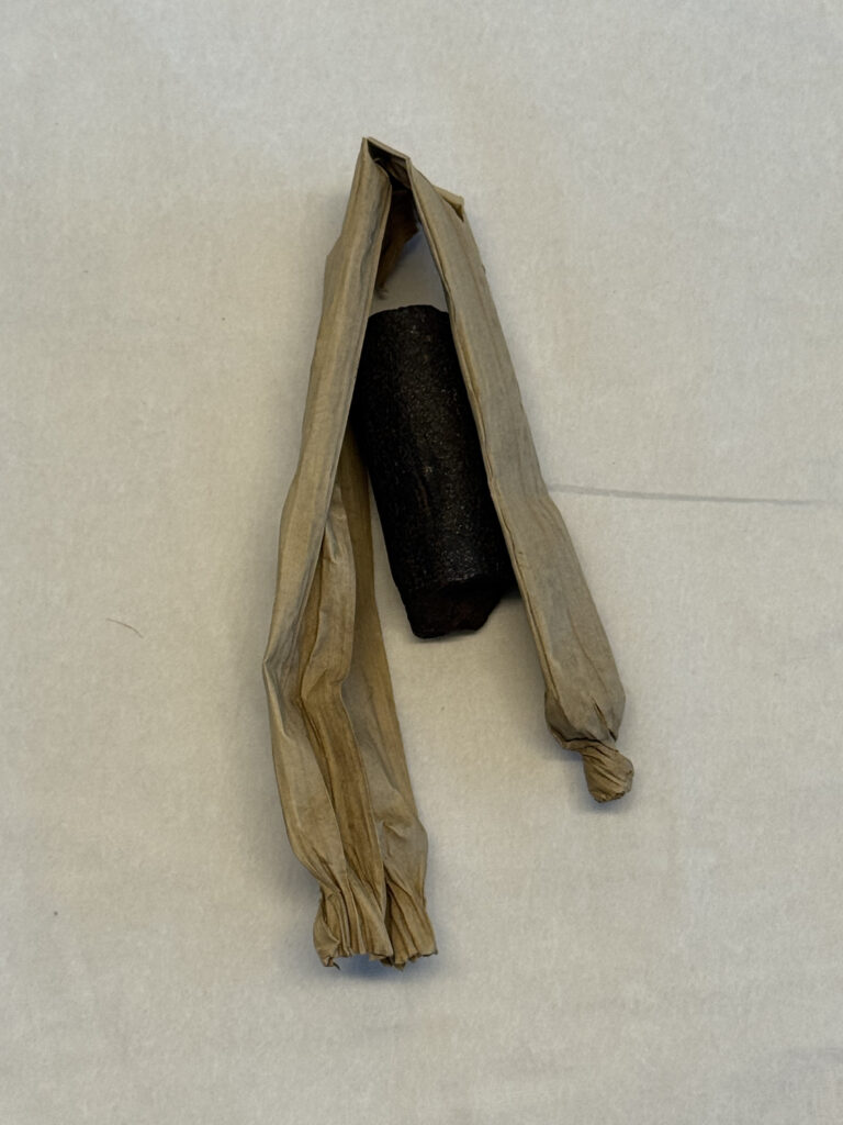 Dark red/ black cylindrical piece of solid matter. It has rough ends resembling a thick, blunt crayon. It has a thin, folded piece of cream paper surrounding it that has been twisted at either end. This acts as the piece's wrapper.
