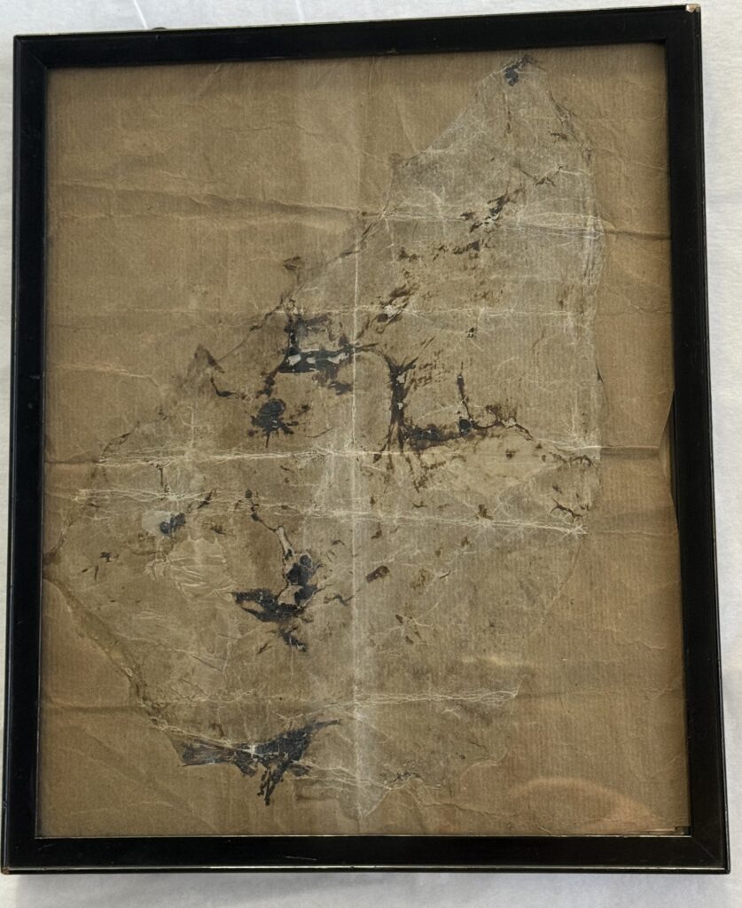 Brown paper with discoloration and patches of black extending across the page in a rough triangular shape. Framed in a black frame.