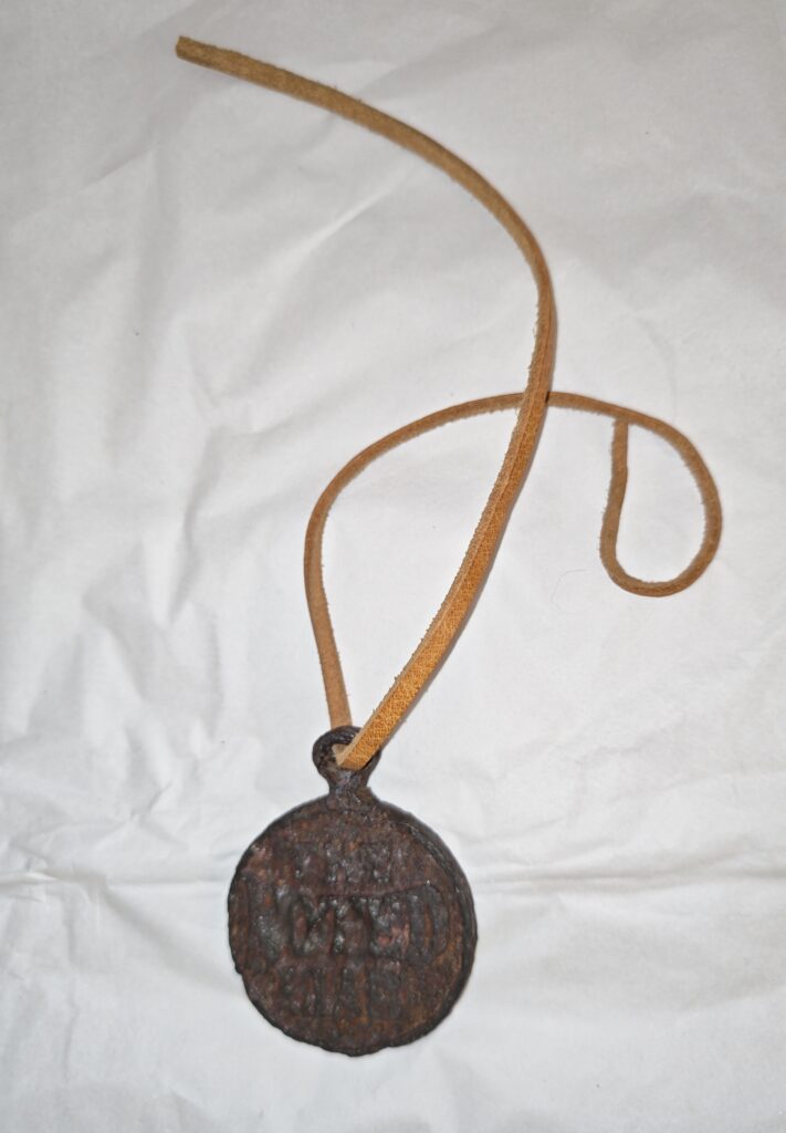 Weathered and rusted black medal with a tan leather strap. The medallion is round and has raised text casted into the metal which writes 'The Noted Liar.'