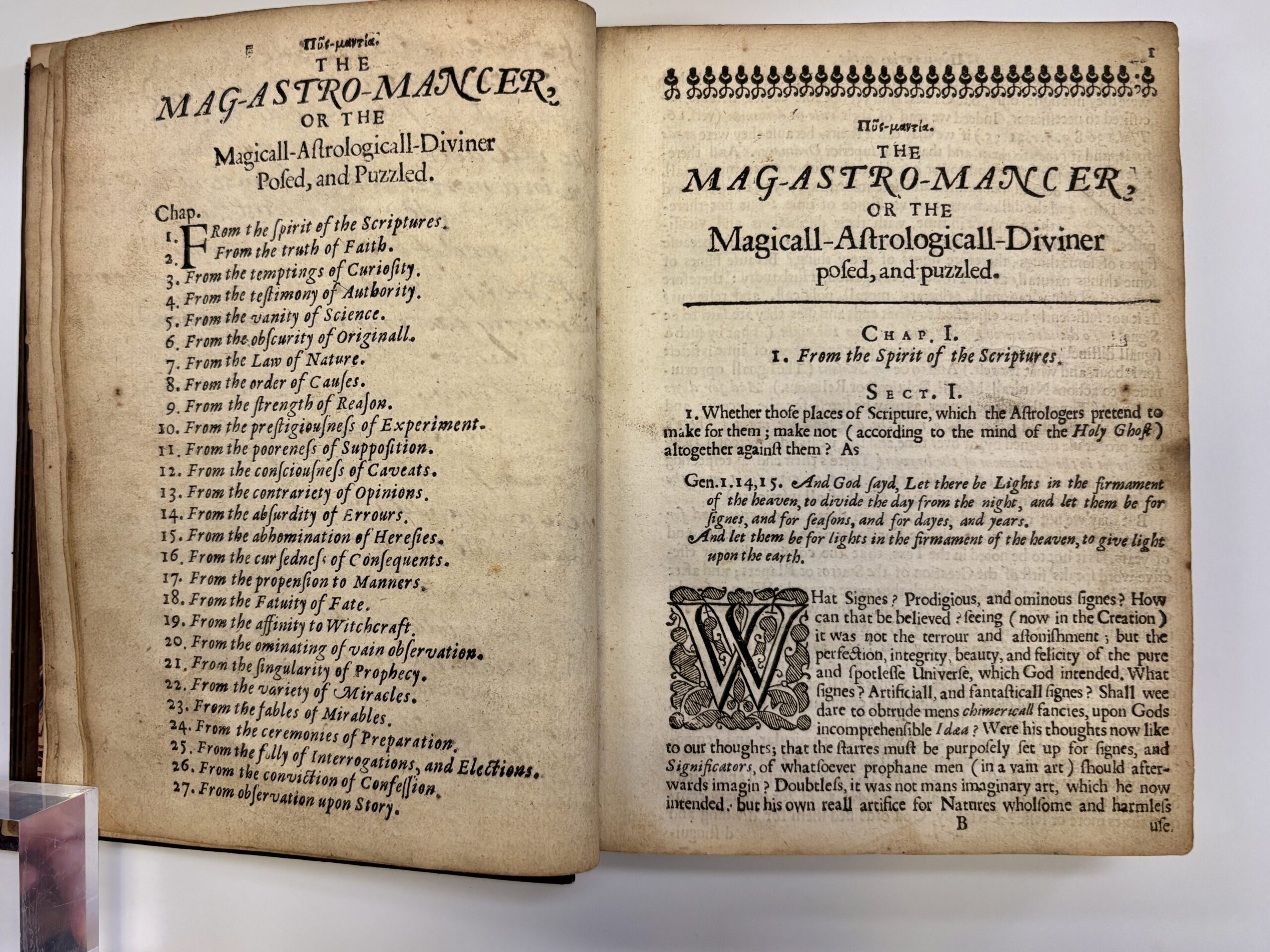 Open book with aged, yellowing pages. Text is in a black 16th-Century typeface, The left-side page reads: 'THE MAG-ASTRO-MANCER OR THE Magicall-Astrologicall-Diviner Posed, and Puzzled.' Beneath this, is a chapter list. 

On the right-side page the page is headed with a black swirling header, the same title is beneath. Below a dividing line is chapter one entitled the 'From the Spirit of the Scriptures'. There is a large, illustrated letter W at the beginning of the paragraph.  