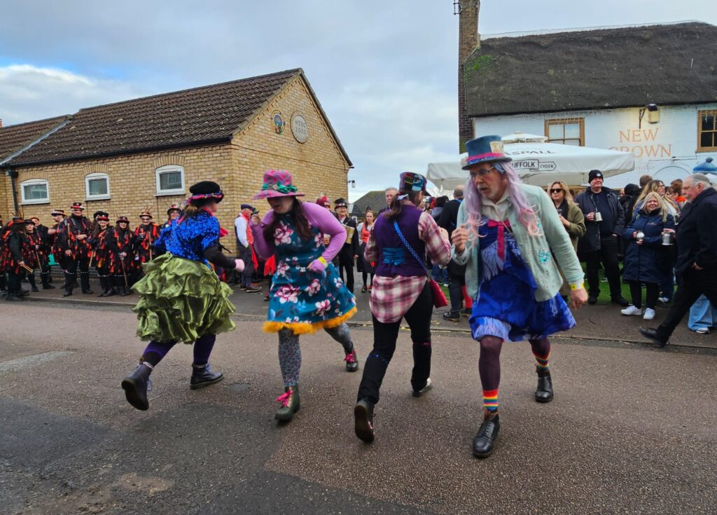 Photograph of Ouse Washes Molly Side, dancing at the Whittlesea Straw Bear Festival, 2026. On a residential street, four people are dancing in colourful clothing and wearing hats with ribbons. A crowd watches on from the pavement. 